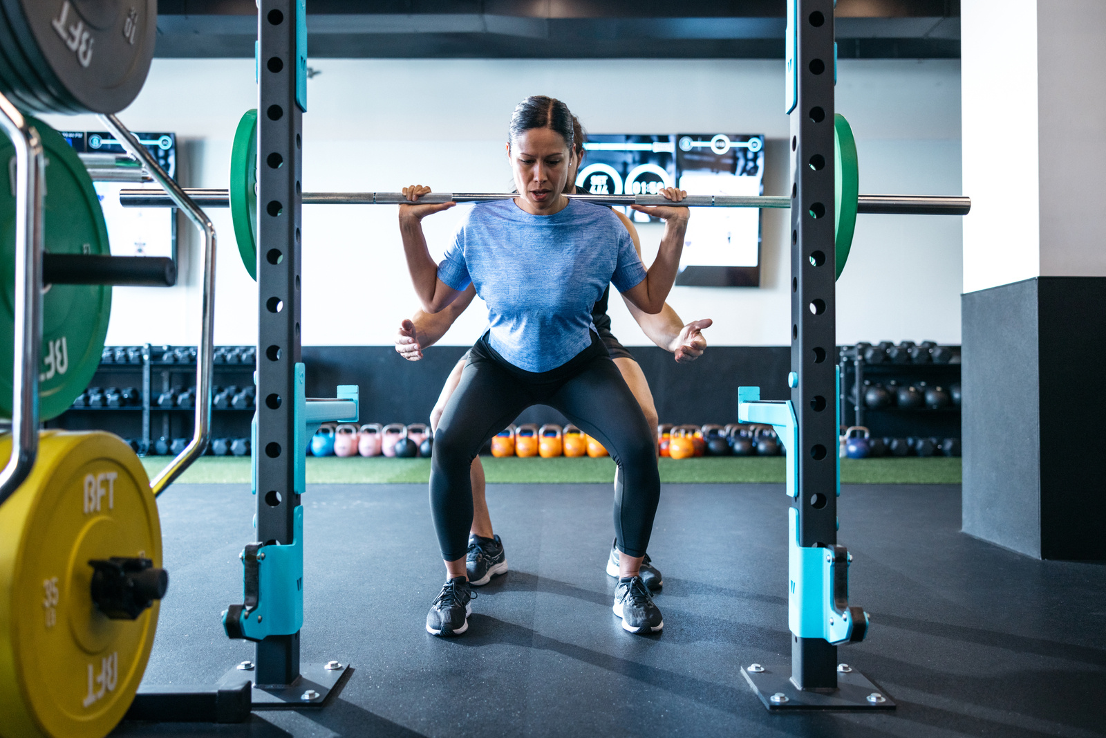 Weight lifting session at a Body Fit Training studio with a female attendee barbell squatting with a trainer assisting.